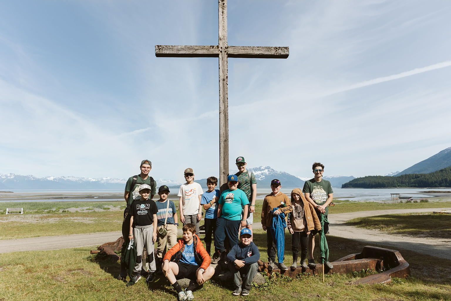 Echo Ranch Bible Campers Group photo of Evan and Echo Ranch campers standing in front of a rustic wooden cross with majestic Alaskan mountains and lake in the background.
